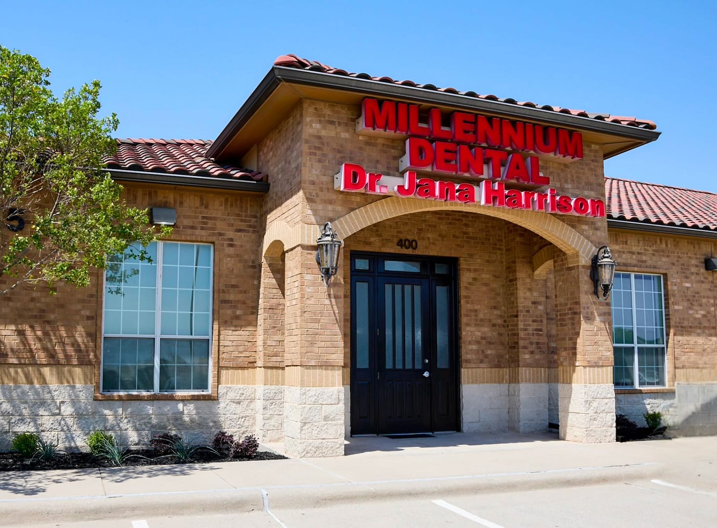 office building made of brown brick and white stone with a spanish tile roof and a sign at the top with Millennium Dental Dr. Jana Harrison in red letters