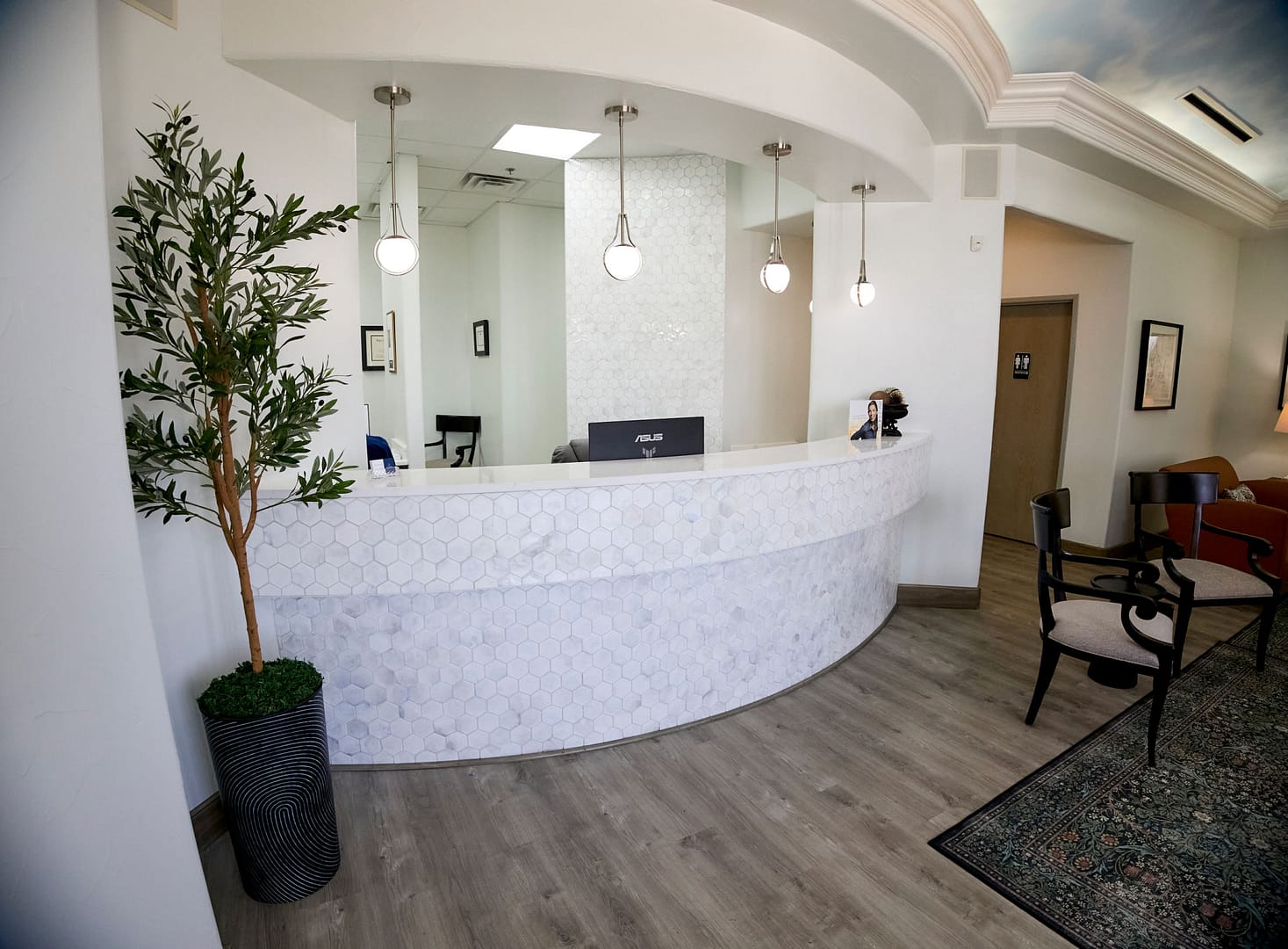 front desk with white and gray marble hexagon tiles on the front and silver pendant lights hanging from the ceiling above it, faux tree in pot on the left