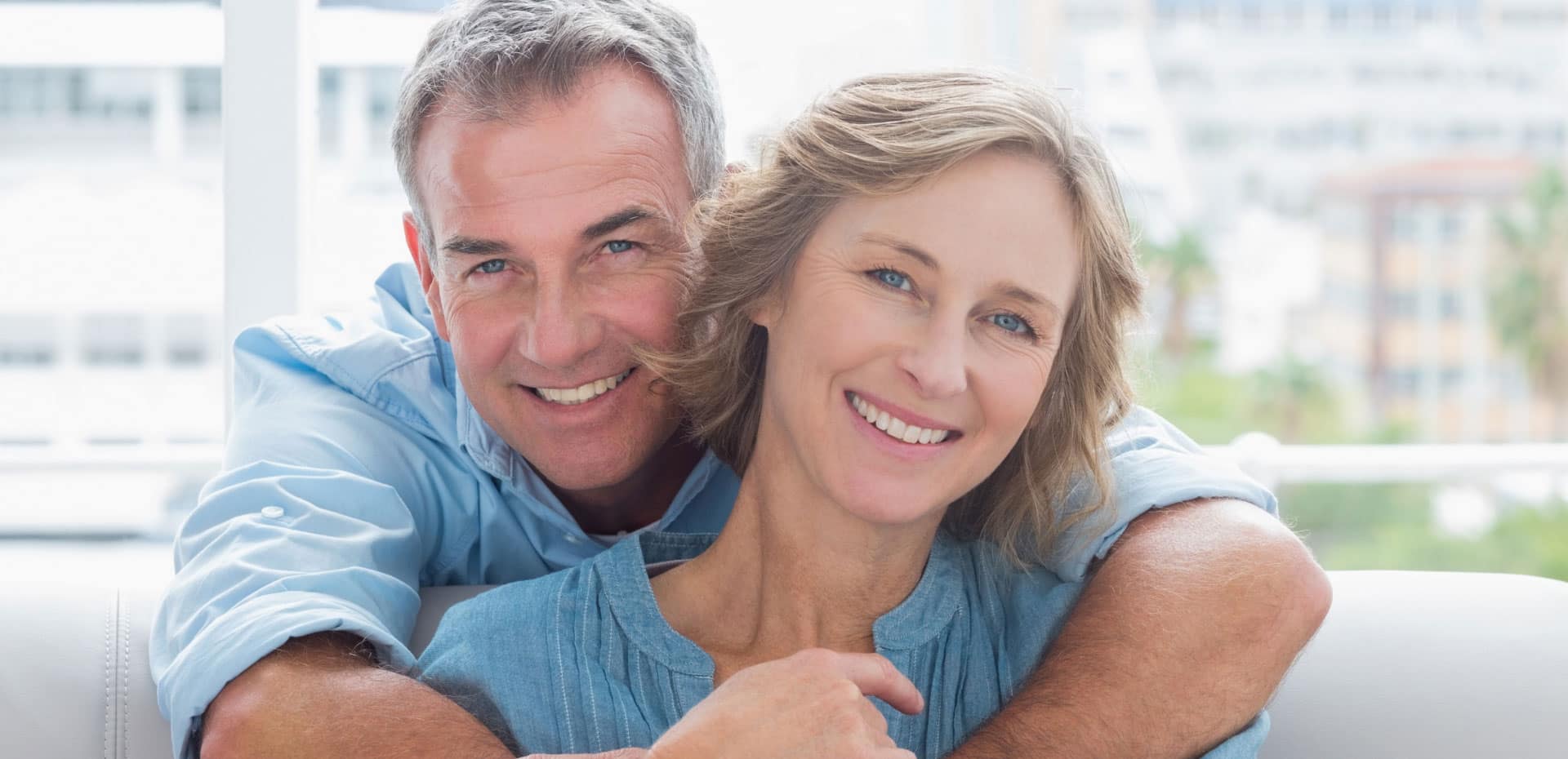middle aged woman sitting on couch while husband leans over couch and hugs her from behind