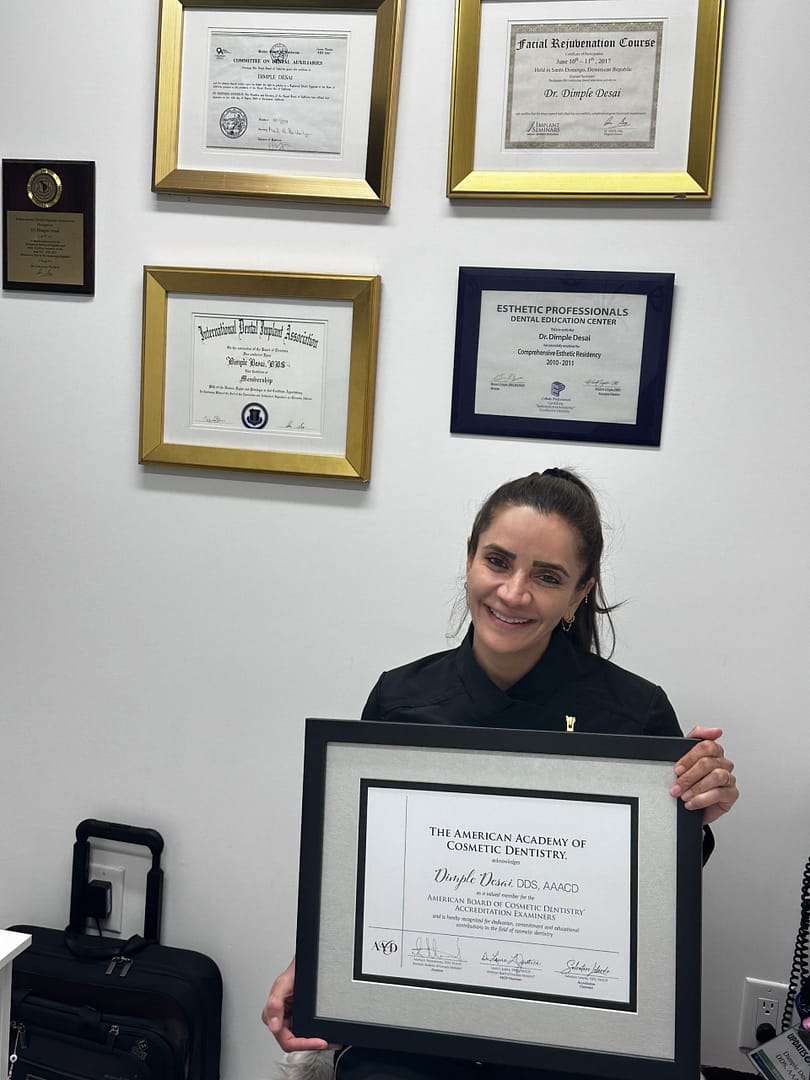 Dr. Desai holding an AACD Accreditation certificate while sitting in front of a wall with other framed certificates